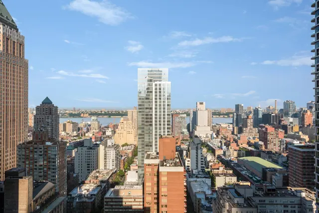 Elevated view over a dense city of mid-rise and high-rise buildings, with a river and distant far shore beneath a partly cloudy blue sky.