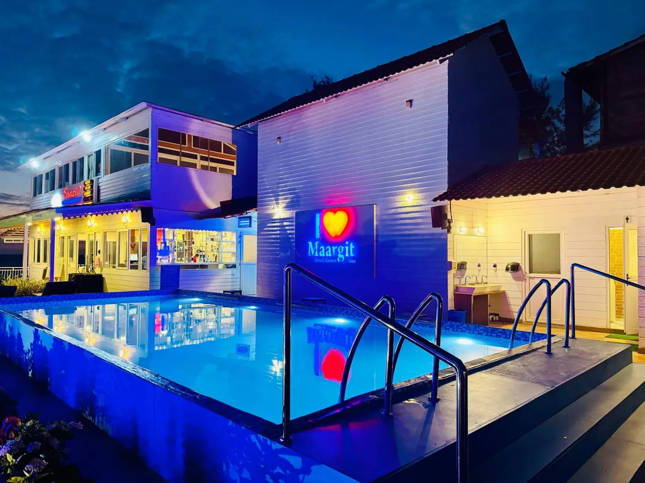 An illuminated rectangular swimming pool stands in front of the hotel buildings at night.