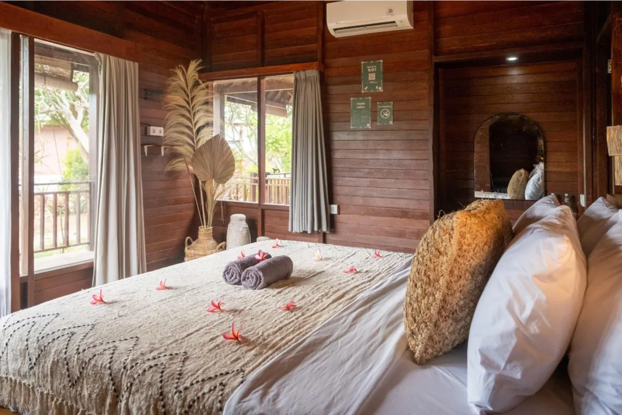 A wooden-walled bedroom with a made bed decorated with frangipani petals and rolled towels, a wall-mounted air conditioner, and windows looking out onto garden trees.