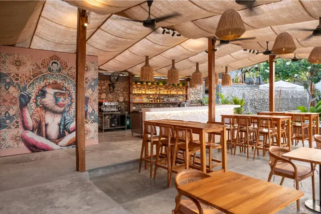 Open-air bar and dining area with a woven canopy roof, rattan pendant lamps, wooden tables and stools, a monkey mural on the left, and a garden with white umbrellas visible outside.