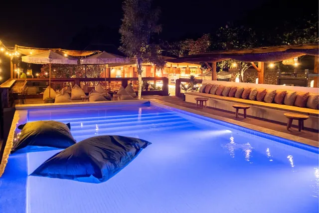 A illuminated blue pool at night with two dark bean bags floating on the water, with lounge seating and fringed umbrellas beyond.