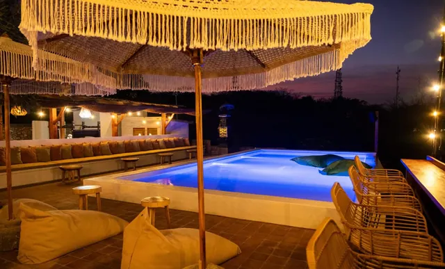 An illuminated pool at night with fringed macrame umbrellas, bean bags, rattan chairs and a long cushioned bench along the wall.
