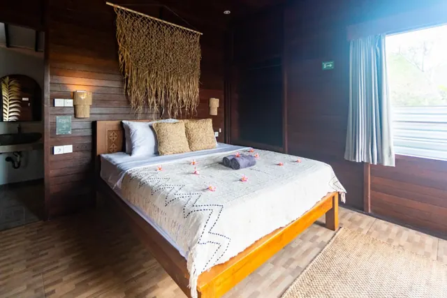 A bedroom with a double bed under a woven throw scattered with petals and two folded towels, a macrame wall hanging, wood-panelled walls, and a window with a curtain showing trees outside.