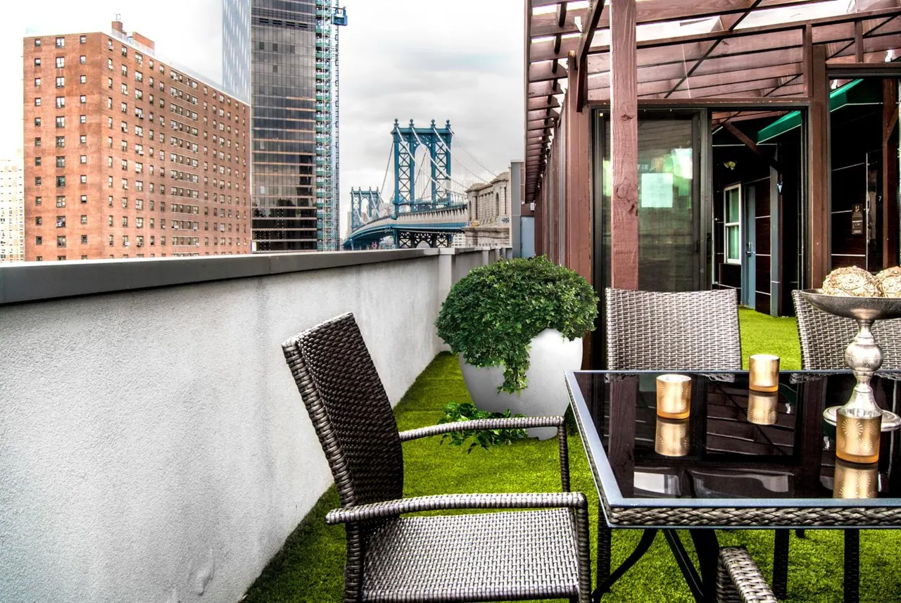 A rooftop terrace with artificial turf, wicker chairs, a glass-top table and a planter, overlooking brick buildings and the Manhattan Bridge.
