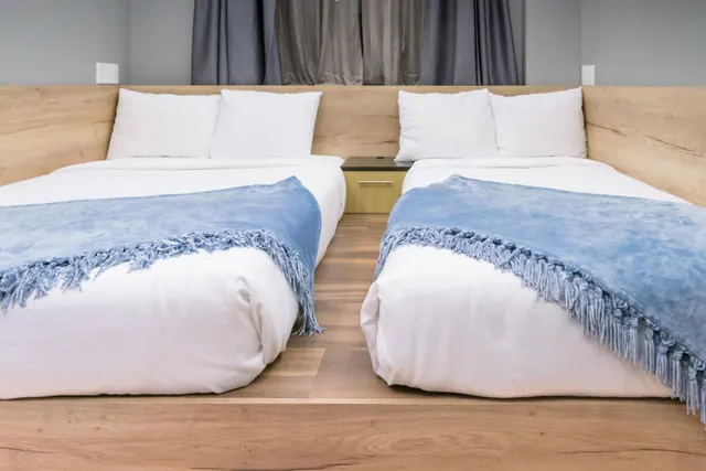 Two made-up double beds with white linen, blue fringed throws and white pillows sit against a wooden headboard wall beside a small wooden nightstand.
