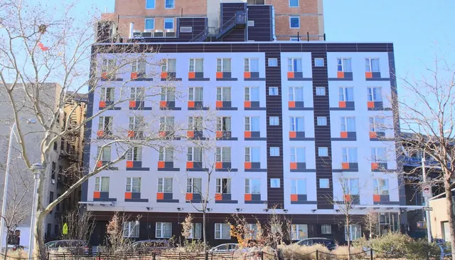A multi-storey white and purple residential-style building with orange window accents stands behind bare winter trees, with parked cars along the street in front.