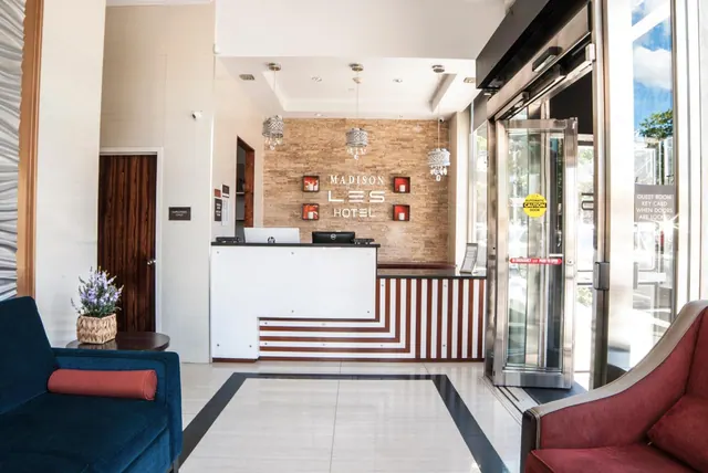 A hotel lobby with a reception desk, a wall sign reading Madison Les Hotel, a blue armchair and a red armchair, and a glass lift.