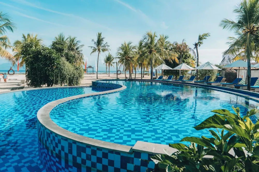 A curved blue-tiled swimming pool with sun loungers and white umbrellas, with a sandy beach, palm trees and the sea beyond.
