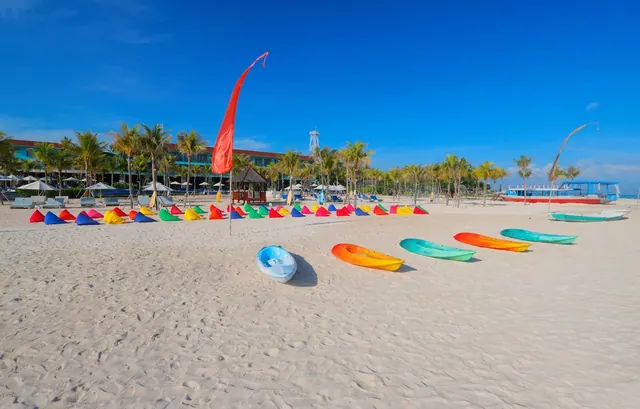 Colourful kayaks and a red flag stand on a sandy beach, with sun loungers, umbrellas and a hotel building behind.