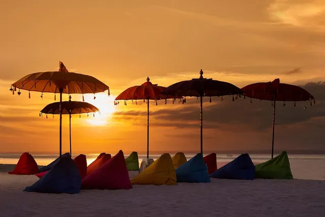 Colourful bean bags and Balinese parasol umbrellas stand on a sandy beach at sunset, with the sea on the horizon.