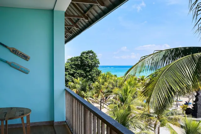 A private balcony with a round wooden table, looking over palm trees toward the sea and a sandy beach.