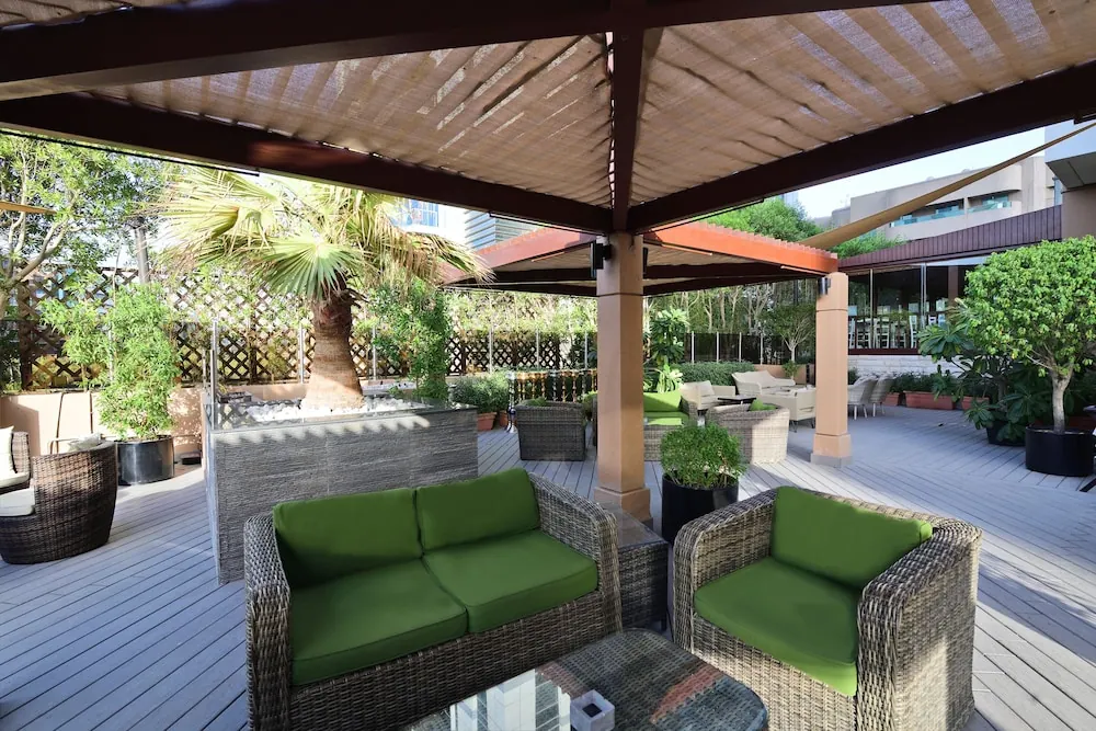 A covered outdoor terrace with green cushioned wicker sofas and armchairs, a palm tree and potted plants under a woven pergola canopy.