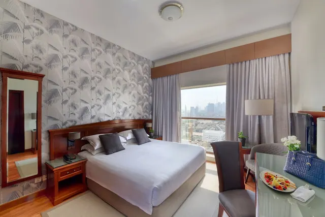 A bedroom with a made king bed, a patterned feature wall, a glass desk with a television, and a window showing a city skyline.