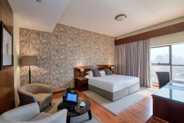 A hotel bedroom with a king bed, two armchairs and a coffee table holding a laptop, with a desk by the window and a city skyline visible outside.
