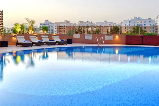 A rooftop swimming pool at dusk with sun loungers with blue towels, potted plants and city buildings visible behind the terrace wall.