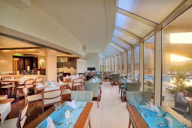 A restaurant dining room with blue-set tables and a glass-walled section looking out over an outdoor pool with sun loungers, where one person sits on the terrace.