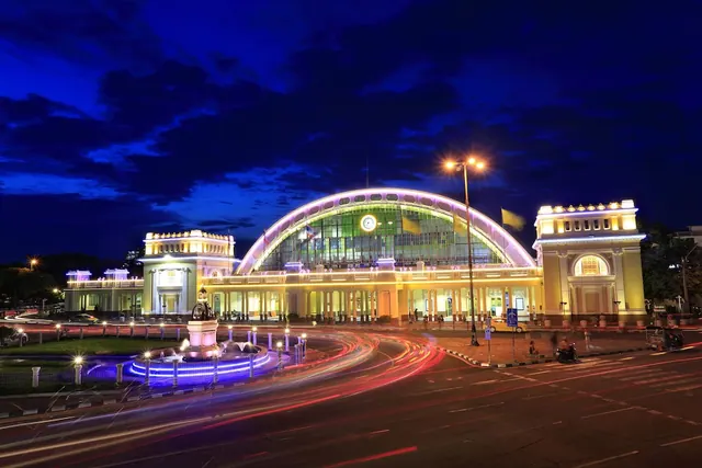 Hua Lamphong Railway Station is lit up at night, with traffic light trails on the road and a illuminated fountain in front.