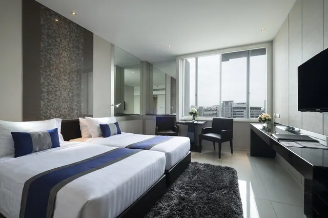 A bedroom with two beds dressed in blue and white linen, a wall-mounted television, armchairs, and a window overlooking city buildings.