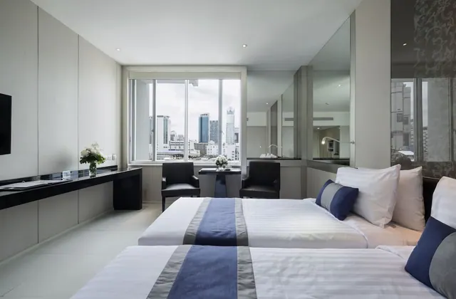 Two twin beds with blue runners sit in a room with a desk, two armchairs and a window overlooking city high-rises.