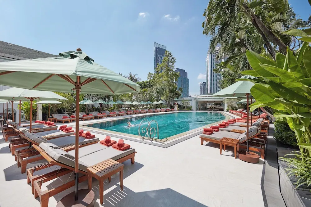 A large outdoor pool with teal umbrellas and sun loungers on the deck, with high-rise towers visible behind the palm trees.