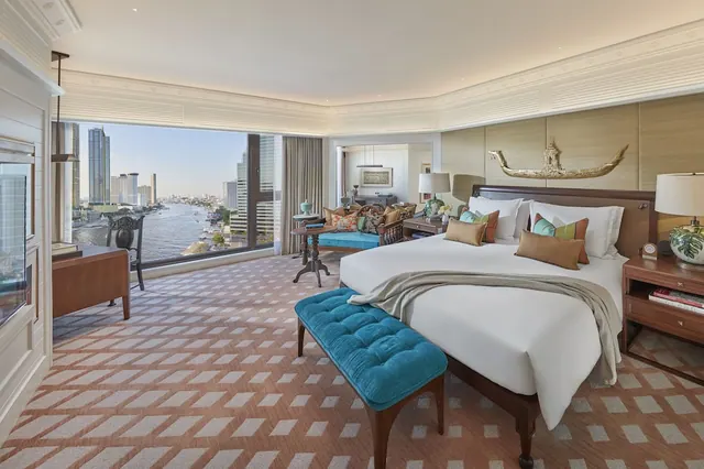 A bedroom with a large white bed, a teal bench at its foot, and floor-to-ceiling windows overlooking a river and city towers.