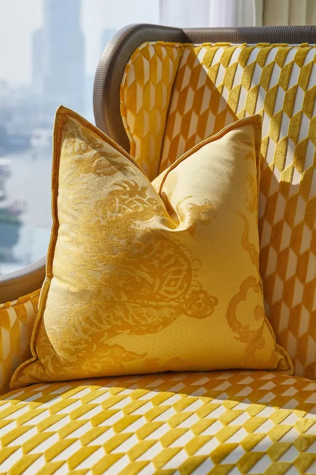 A yellow geometric-patterned armchair with an embroidered gold cushion sits beside a window showing a river and high-rise buildings.