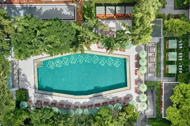Aerial view of an outdoor swimming pool with turquoise water and a green patterned tile border, surrounded by sun loungers, green parasols and tropical trees.