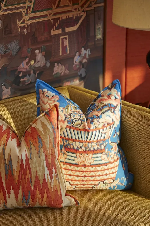 Two decorative cushions, one with a blue floral print and one with an orange ikat pattern, sit on a mustard sofa in front of a painted Thai-style mural and a lamp.