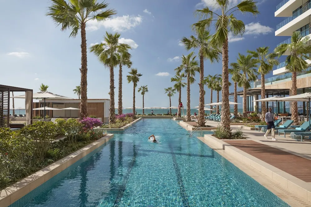 A long lap pool lined with palm trees and planting, with sun loungers and white umbrellas nearby, a swimmer in the water, and the sea and beach visible in the distance.
