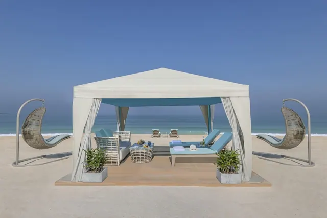 A white canopied beach cabana with blue daybeds, a wicker sofa and two hanging wicker chairs stands on sand facing the sea.