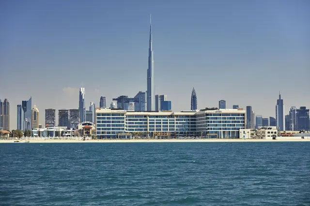 A low-rise hotel building stands on a sandy beach facing the sea, with the Burj Khalifa and other skyscrapers rising behind it.