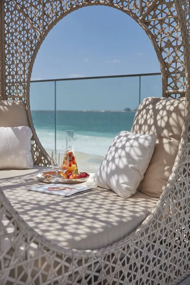 A white wicker daybed with cushions sits on a terrace overlooking the sea, with a jug of fruit drink, glasses, and a magazine on a table.