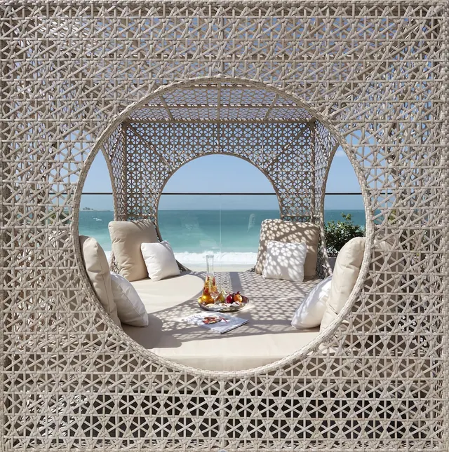 A woven daybed with cushions, a carafe of drink and a fruit tray sits in an open cabana framed by a view of the sea and breaking waves on the shore.