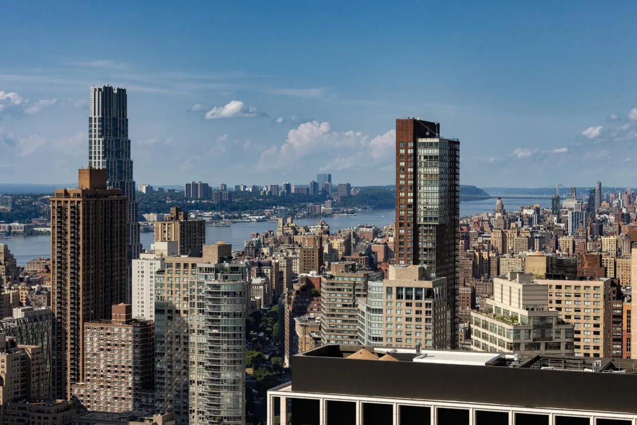 Elevated view over Manhattan high-rises toward the Hudson River and the wooded far shore under a partly cloudy blue sky.