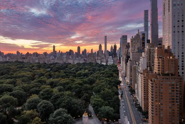 Aerial view over Central Park toward a city skyline at sunset, with tall residential towers on the right and a long avenue running between them.