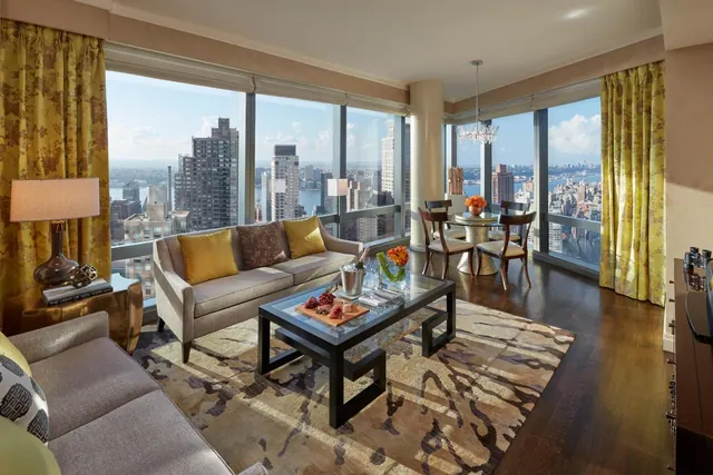 A living room with a grey sofa, a glass coffee table and a round dining table beside floor-to-ceiling windows overlooking city buildings and a river.