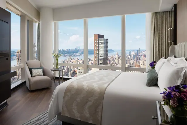 A bedroom with a made king bed, an armchair, and floor-to-ceiling windows looking out over a city skyline and a river.