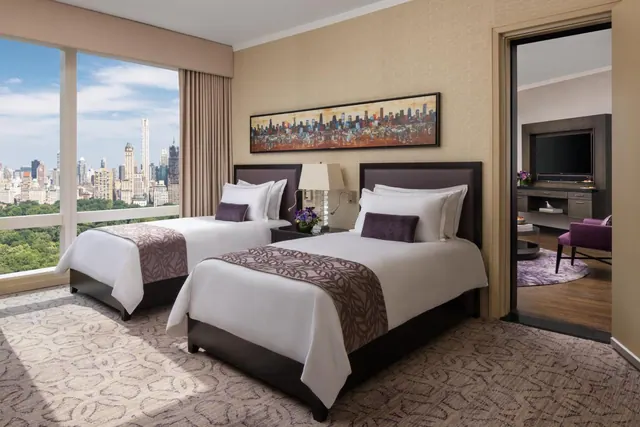 A bedroom with two twin beds beside floor-to-ceiling windows overlooking a park and city skyline, with an adjoining room with a television visible through the open doorway.