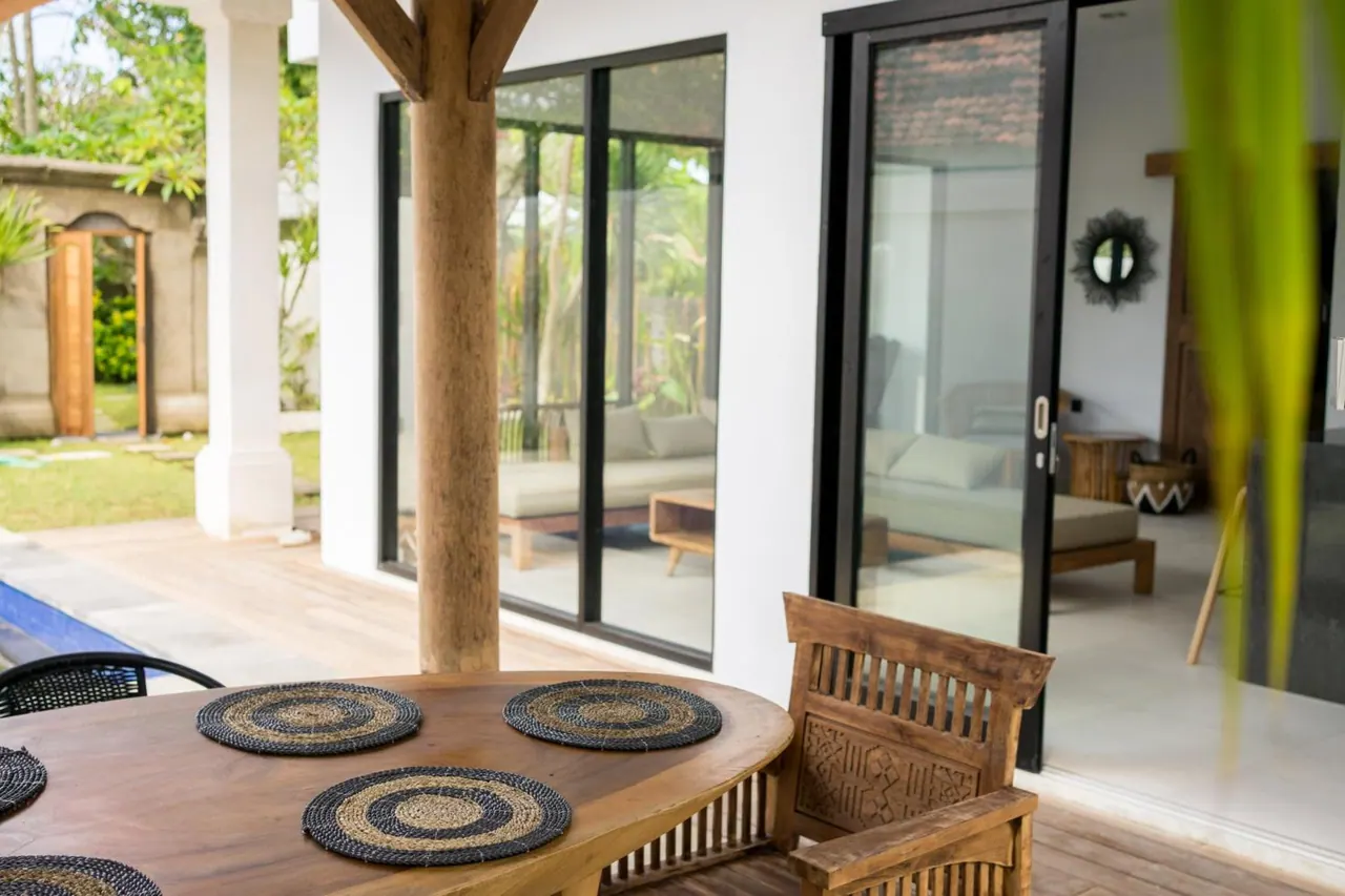 A covered terrace with a round wooden dining table, woven placemats and a carved wooden chair beside glass doors that open into a living room, with a garden visible through the windows.