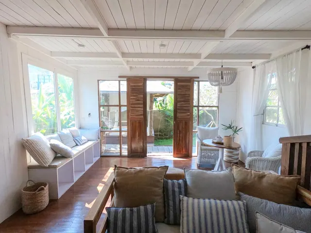A bright living area with a white timber ceiling, a built-in window seat with cushions, a sofa in the foreground, and glass doors opening onto a garden.