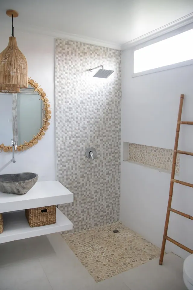 A walk-in shower with mosaic wall tiles and a pebble floor, beside a stone basin on a floating white vanity and a round rattan mirror.