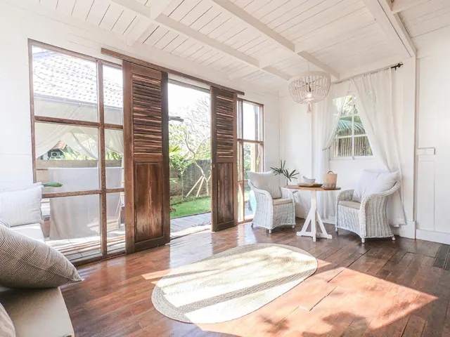 A bright white room with dark wood floors, wicker armchairs, a small round table and an oval rug, with glass doors open onto a garden.