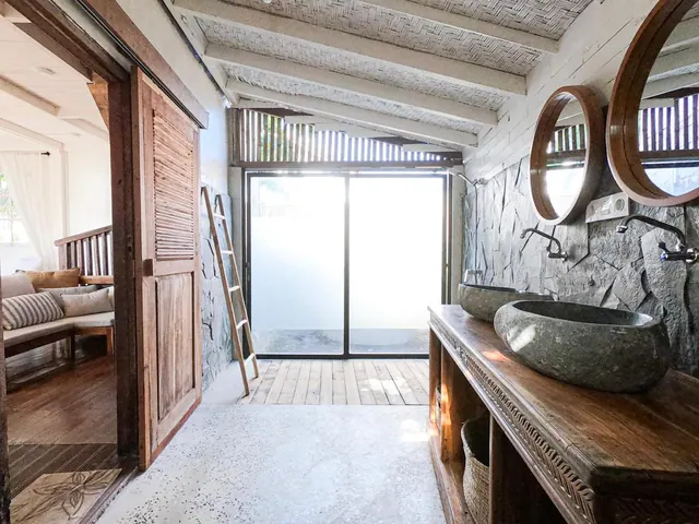 A bathroom with two stone basins on a wooden vanity, round wooden mirrors, a wooden ladder, and sliding glass doors to an outdoor area, with a seating area visible through the doorway on the left.