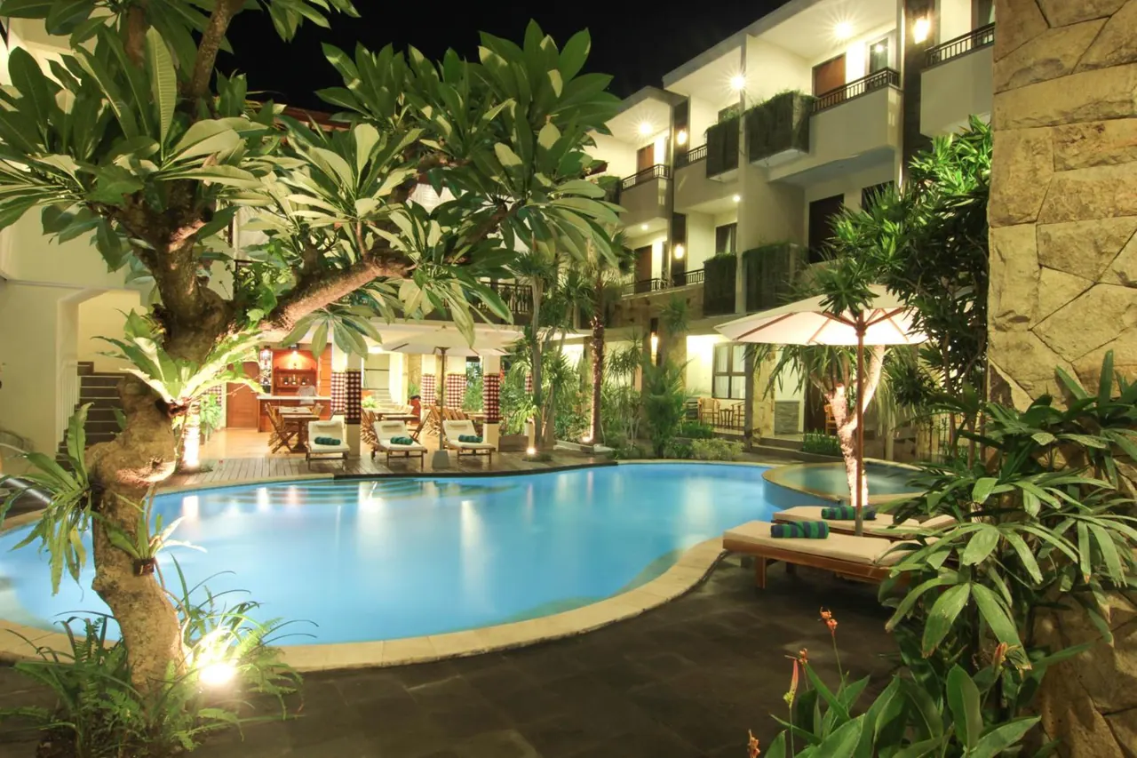 A illuminated freeform swimming pool at night with sun loungers and a white parasol beside it, with a frangipani tree on the left and hotel balconies behind.