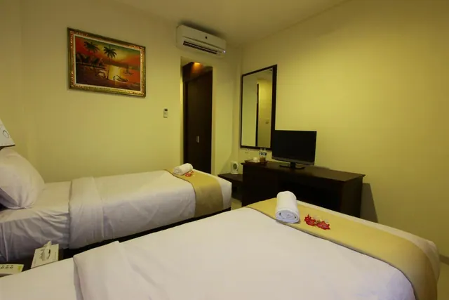 A hotel bedroom with two single beds dressed with towels and red flower decorations, a wall-mounted air conditioner, a framed painting, and a flat-screen television on a wooden desk.