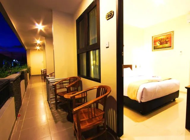 A covered outdoor walkway at dusk with wooden chairs and a small table, and an open door showing a bedroom with a bed and a framed painting.