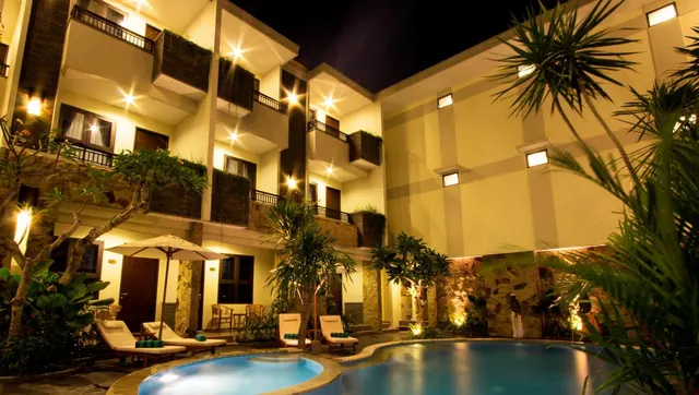 A illuminated hotel pool in a courtyard at night, with sun loungers, a white umbrella and balconies on the surrounding building.