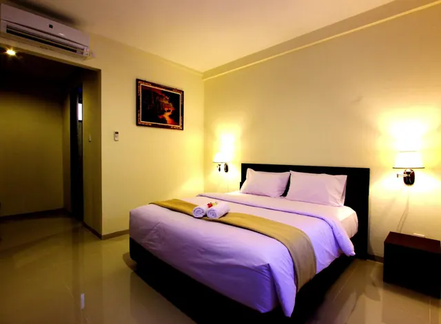 A double bed with lavender bedding and rolled towels on a tan runner sits between two lit wall lamps, with a wall-mounted air conditioner near the ceiling at left.