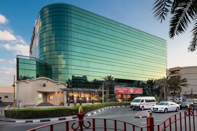 Exterior view of the Marco Polo Hotel with its curved green glass facade, entrance canopy, and cars on the driveway.