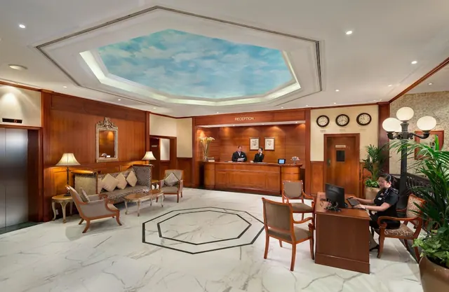 A hotel lobby with a marble floor, a wood-panelled reception desk with two staff behind it, seating groups, a lift on the left, and a painted sky panel in the ceiling.